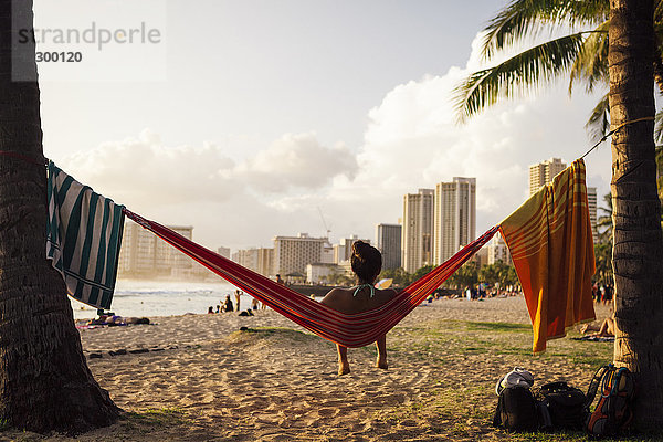 Ein spanischer Erwachsener entspannt sich in einer Hängematte am Waikiki Beach  Hawaii  während die Sonne an einem warmen Sommerabend untergeht.