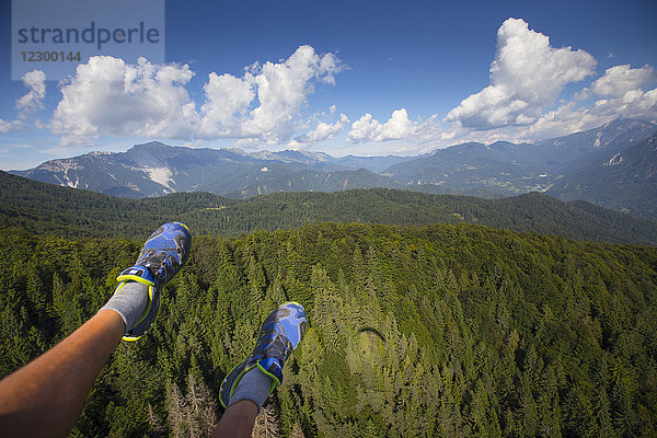 Die Füße eines Gleitschirmfliegers baumeln hoch über den Wäldern der slowenischen Alpen während eines Gleitschirmfluges vom Berg Dobrca bei Begunje und Radovljica in Slowenien. Auf den Baumwipfeln ist ein Schatten zu sehen.