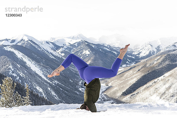 Junge Frau macht Kopfstand auf einem verschneiten Hügel vor einem Bergtal  Aspen  Colorado  USA