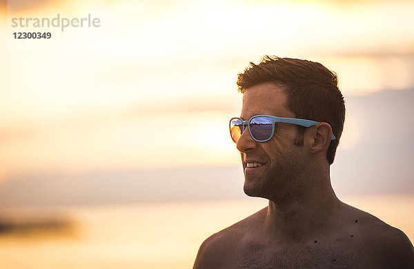 Lächelnder Mann ohne Hemd mit Sonnenbrille bei Sonnenaufgang