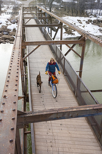 Ein Hund läuft neben einer erwachsenen Frau  die mit dem Fahrrad über eine Brücke über den Animas River fährt  Durango  Colorado  USA