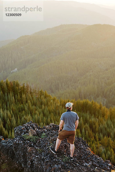 Wanderer mit Kamera auf dem Gipfel des Iron Mountain in Oregon. Dieser Wanderer steht auf dem Gipfel einer 700 Fuß hohen Klippe und überblickt einen Großteil des Willamette National Forest in Oregon. Aufgenommen bei Sonnenuntergang mit schönem Gegenlicht und einige dunstigen Himmel von Waldbränden  die Oregon während des Sommers 2017 geplagt.
