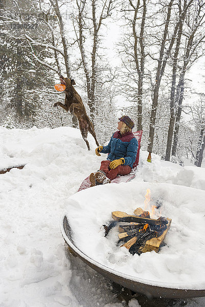 Frau entspannt sich im Schnee an einer brennenden Feuerstelle und spielt mit einem Hund Plastikring  Durango  Colorado  USA