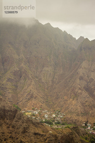 Wolken umhüllen den Gipfel eines Berges auf den Kapverden  Afrika  während sich ein kleines Dorf ruhig in ein Tal darunter schmiegt.