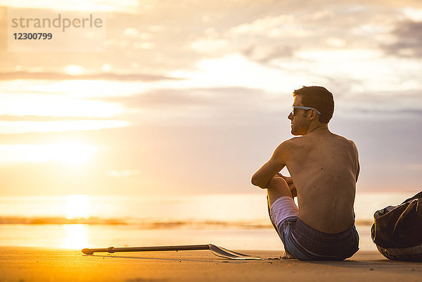 Rückansicht eines einsamen Mannes ohne Hemd  der bei Sonnenaufgang am Strand sitzt