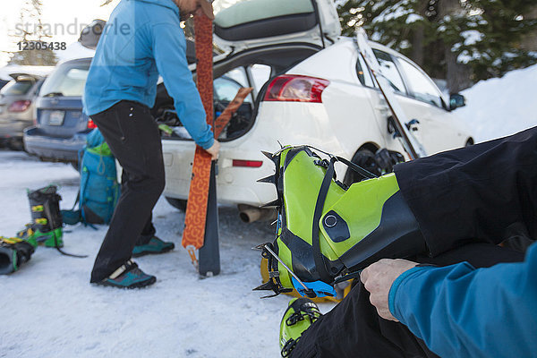 Zwei Skitourengeher bereiten sich auf das Skifahren vor  wobei einer die Skischuhe anzieht und der andere den Kofferraum des Autos erreicht
