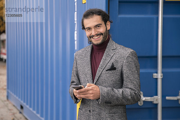 Lächelnder Hispanoamerikaner mit Blazer und Bart  der neben einem Container steht und sein Mobiltelefon benutzt