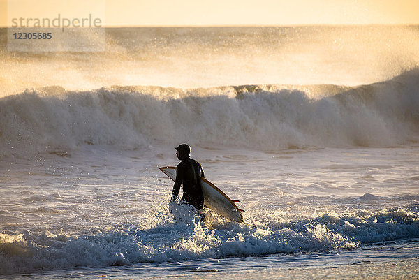 Surfer im Neoprenanzug geht und trägt Surfbrett im Meer bei Sonnenuntergang