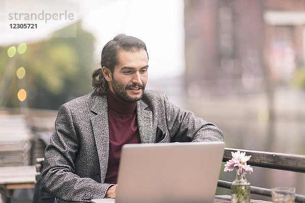 Hispanischer Mann mit Bart  der draußen in einem Café am Wasser einen Laptop benutzt und einen Blazer und einen Rollkragenpullover trägt