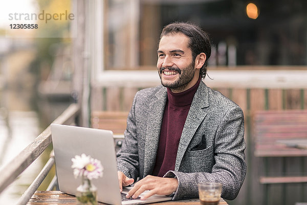 Hispanischer Mann mit Bart  der draußen in einem Café am Wasser einen Laptop benutzt und einen Blazer und einen Rollkragenpullover trägt