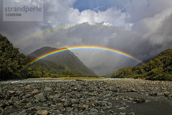 Doppelter Regenbogen vor großen Wolken über felsigem Tal  Neuseeland