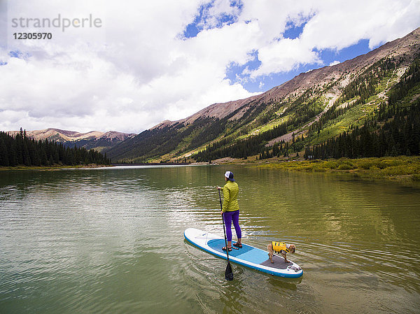 Frau paddelt mit ihrem Hund auf einem Paddelbrett auf einem Stausee in den Bergen bei Aspen  Colorado  USA