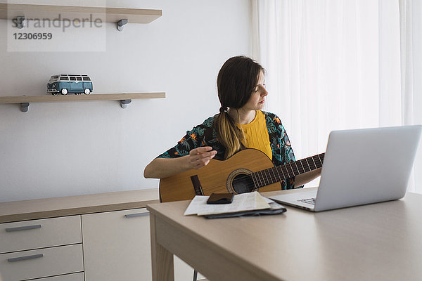 Junge Frau sitzt zu Hause am Tisch und spielt mit dem Laptop Gitarre.
