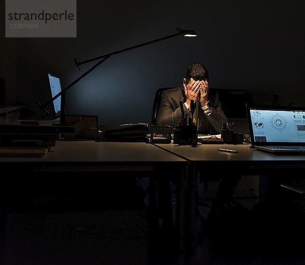 Überlasteter Geschäftsmann sitzt an seinem Schreibtisch im Dunkeln