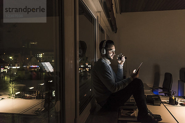 Geschäftsmann sitzt nachts auf der Fensterbank im Büro und hört Musik mit Kopfhörern.