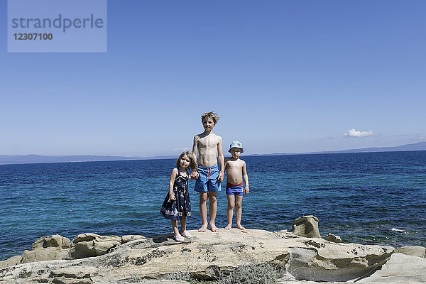 Griechenland  Chalkidiki  drei Kinder  die Hand in Hand auf Felsen vor dem Meer stampfen