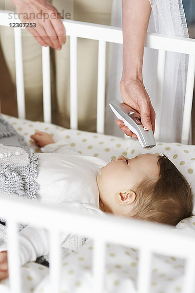Mutter mit digitalem Thermometer  das die Temperatur des Babys in der Krippe überprüft.
