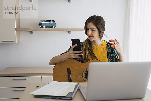 Junge Frau zu Hause am Tisch sitzend mit Gitarre  Handy und Laptop