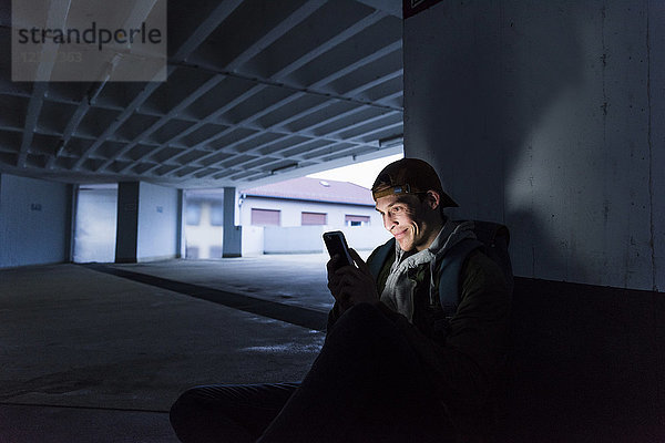 Lächelnder Mann schaut auf leuchtendes Smartphone im Parkhaus