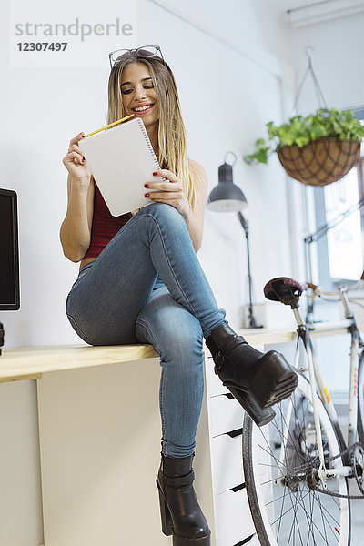 Lächelnde junge Frau sitzt auf dem Schreibtisch im Büro und macht sich Notizen.