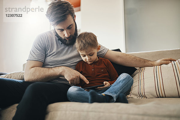 Vater und kleiner Sohn sitzen zusammen auf der Couch und schauen auf das Smartphone.