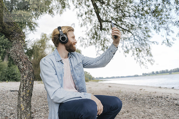 Deutschland  Düsseldorf  Mann mit Kopfhörer nimmt Selfie am Strand mit Smartphone