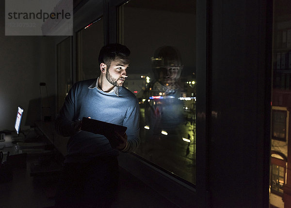 Geschäftsmann sitzend mit Tablette auf Fensterbank im Büro bei Nacht aus dem Fenster schauend