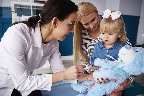 Arzt und Mädchen mit Mutter impfender Teddy in der Arztpraxis