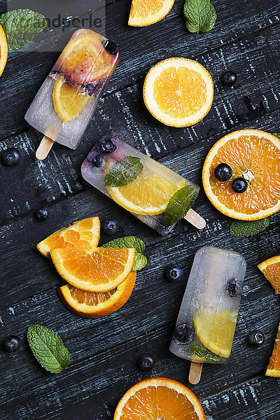 Hausgemachte Entgiftungs-Popsicles mit Heidelbeeren  Orangenscheiben und Minzeblättern auf schwarzem Holz