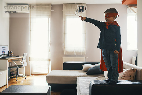 Kleiner Junge verkleidet als Superheld  der zu Hause auf einem Couchtisch steht und auf etwas zeigt.