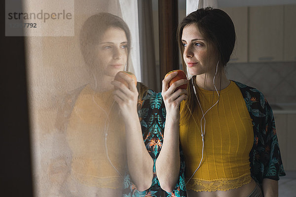 Junge Frau mit Kopfhörer und Apfel aus dem Fenster schauend
