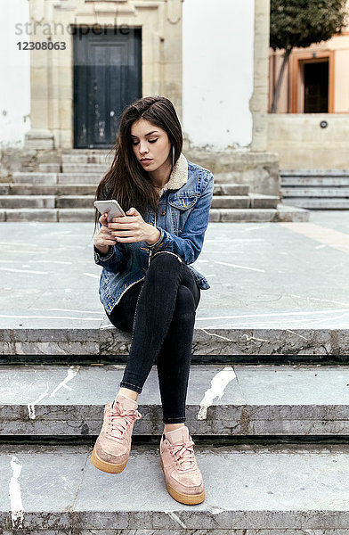Junge Frau sitzt auf einer Treppe in einer Stadt und überprüft ihr Smartphone.