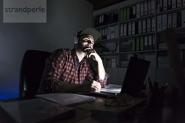 Porträt eines Freiberuflers  der nachts am Schreibtisch sitzt  mit Laptop und Kopfhörer