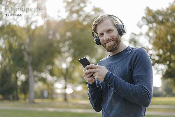 Porträt eines lächelnden Mannes mit Handy beim Musikhören mit Kopfhörern im Park