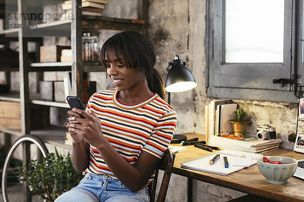 Lächelnde junge Frau sitzt vor dem Schreibtisch in einem Loft und schaut auf das Handy.