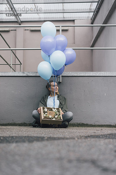Lächelnde Frau mit Blumen in Pappschachtel und blauen Luftballons auf dem Boden sitzend