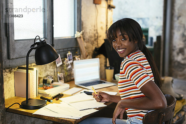 Porträt einer lachenden jungen Frau  die am Schreibtisch in einem Loft sitzt.