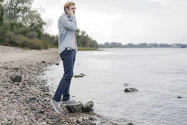 Deutschland  Düsseldorf  Mann am Telefon  der am Flussufer steht und die Aussicht betrachtet