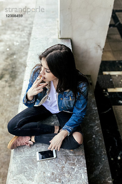 Junge Frau sitzt auf einer Wand und raucht eine Zigarette.