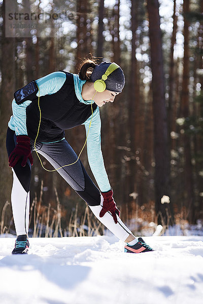 Junge Frau mit Kopfhörer beim Stretching im Winterwald