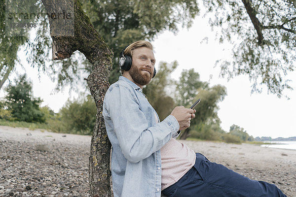 Deutschland  Düsseldorf  Mann beim Musikhören mit Smartphone und Kopfhörer am Strand
