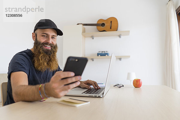 Porträt eines bärtigen jungen Mannes am Schreibtisch mit Laptop beim Blick auf das Handy