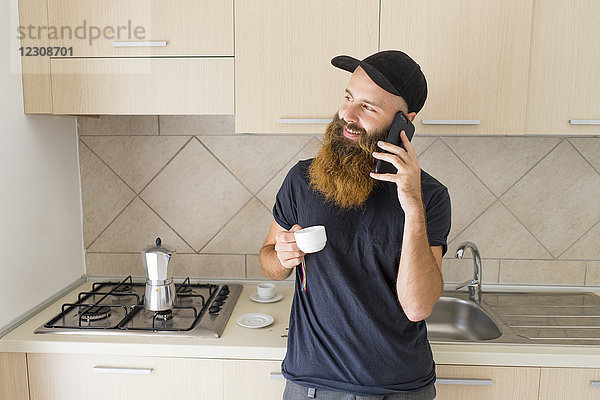 Porträt eines bärtigen Mannes am Telefon stehend in der Küche mit Espressotasse