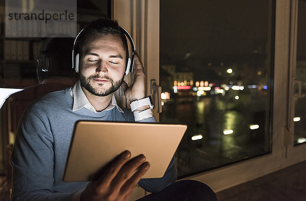 Geschäftsmann sitzt nachts auf der Fensterbank im Büro und hört Musik mit Kopfhörer und Tablett.