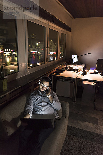 Geschäftsmann im Büro sitzt nachts auf der Couch und benutzt Laptop und Kopfhörer.