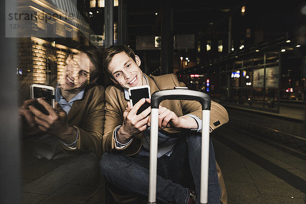 Lächelnder Geschäftsmann mit Handy an der Straßenbahnstation bei Nacht