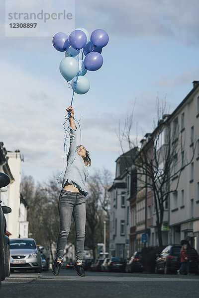 Junge Frau mit blauen Luftballons  die auf der Straße in die Luft springen.