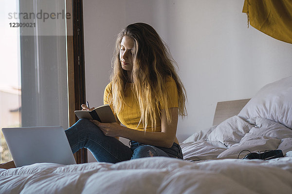 Junge Frau mit Laptop auf dem Bett sitzend  etwas aufschreibend