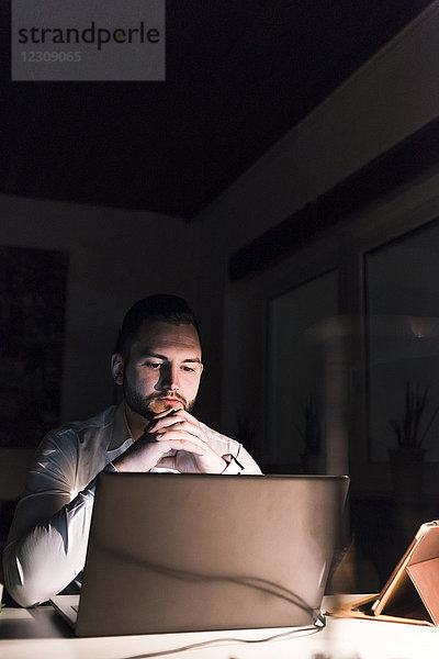 Portrait eines Geschäftsmannes am Schreibtisch im Büro bei Nacht mit Blick auf den Laptop