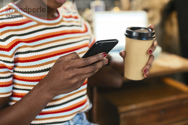 Frau hält Kaffee zum Mitnehmen während der Benutzung des Smartphones  Nahaufnahme